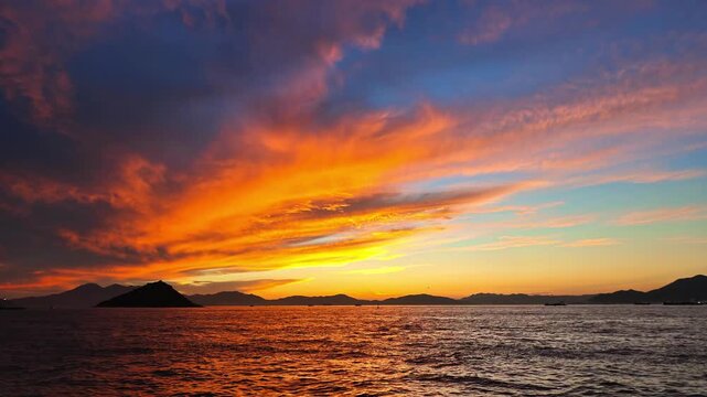 Breathtaking twilight video capturing vibrant orange, red, and blue fiery clouds over a dark sea with a small silhouette island.
