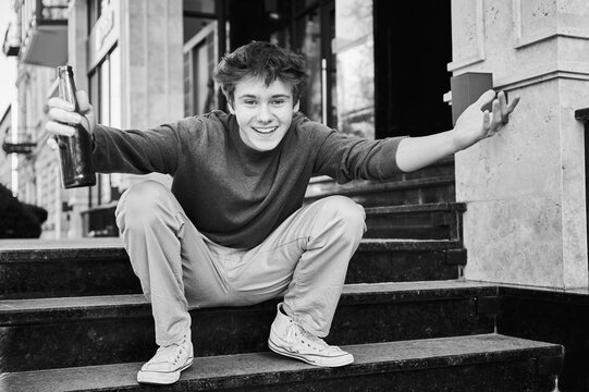 Teen boy sits on city steps holding bottle by knee. Emotional urban scene suggests sadness, stress, and risky behavior. Concept of underage alcohol problem
