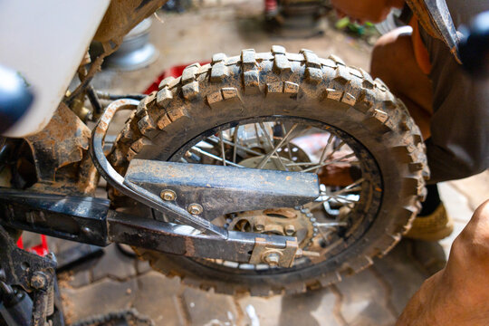 Dirty knobby motorcycle tire on spoked wheel with chain and swingarm being repaired in workshop, gritty detail and hands on mechanical work atmosphere