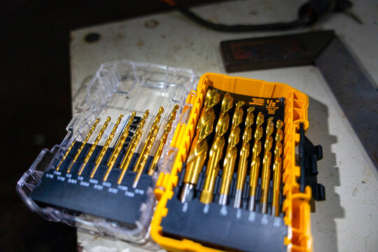 Golden drill bit set in plastic case on workbench with warm light and shallow depth of field showing organized metal tools and workshop atmosphere