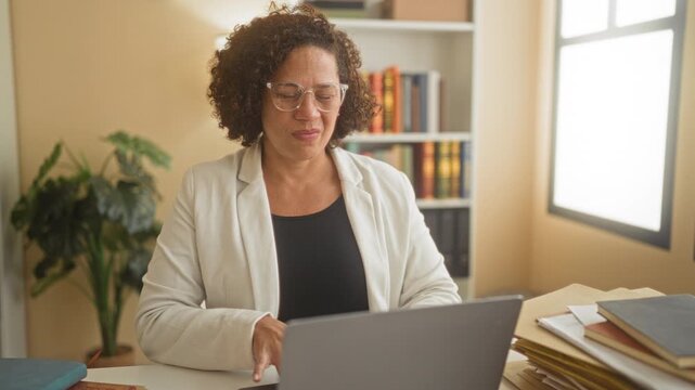 Woman rubbing wrist with a pained expression at laptop on desk in building office, wearing glasses with bookshelf and plant nearby; pain strain.