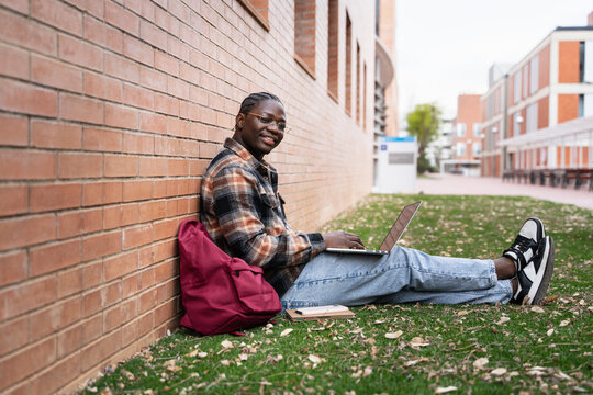 African University student with glasses using laptop sitting on grass at Campus park, looking at camera