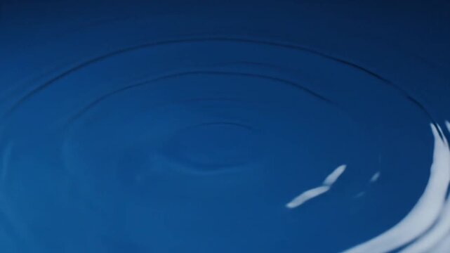 A single water droplet falls into a calm pool of deep blue liquid, creating a crown-like splash followed by expanding concentric ripples that gradually dissipate across the surface.