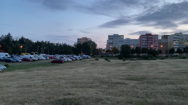 Suburban city edge at summer dusk with multistory apartment buildings and parking area surrounded by open fields. Darkening evening sky creates a calm urban atmosphere. Parking cars
