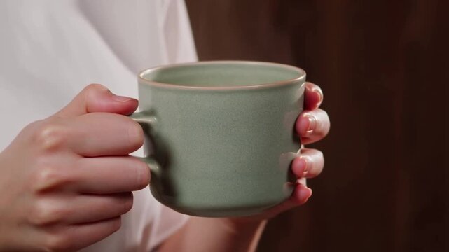 A person holding a steaming green mug of hot coffee or tea against a dark wood background, wearing a white garment.