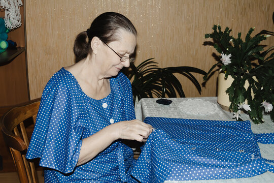 An elderly woman in blue dress focuses intently sewing garment table quiet domestic scene highlighting craftsmanship, patience, and the timeless joy creating clothing hand cozy familiar home setting