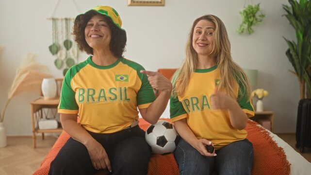 Woman friends sitting on bed points finger to brazil badge on their shirts and holds a soccer ball between them in building; celebration camaraderie.
