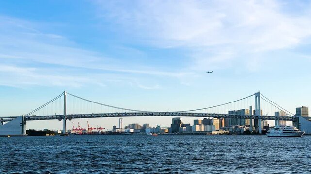 青空のもとレインボーブリッジの全景と東京湾を航行するクルーズ船、上空を通過する旅客機が交差するウォーターフロント景観。