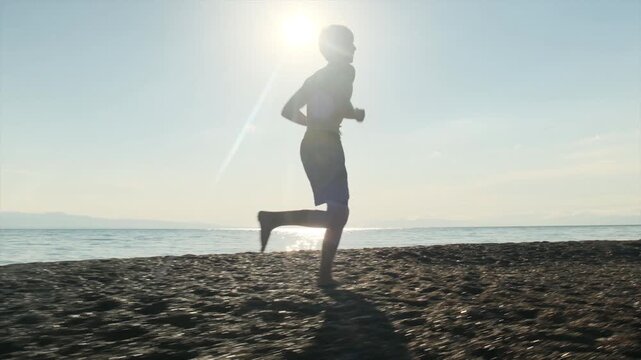 Determined shirtless young athlete runs barefoot along a rocky seashore at sunset. Morning run, healthy lifestyle, endurance, resilience, solo workout, striving for success in life.