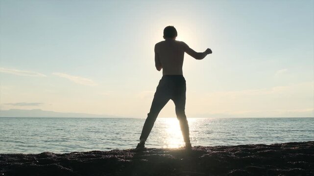 Tall slim muscular young man practices hand to hand combat strikes on the seashore with a scenic sea horizon. MMA athlete, martial arts training, healthy lifestyle, self defense for protecting himself