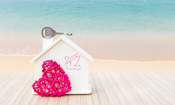 Miniature house with pink heart and house key over tropical beach background, love and romance, valentine background