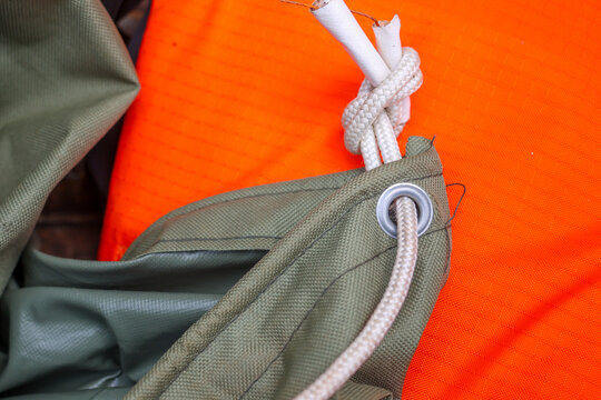 Olive green ripstop tarp corner with metal grommet and white rope knot on bright orange fabric, close up texture detail showing stitching, outdoor gear utility and rugged material