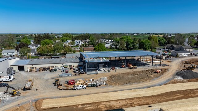 Workers are busy at a construction site in a rural area. Heavy machinery is present, and materials are stacked nearby. The building frame is taking shape under a clear blue sky.