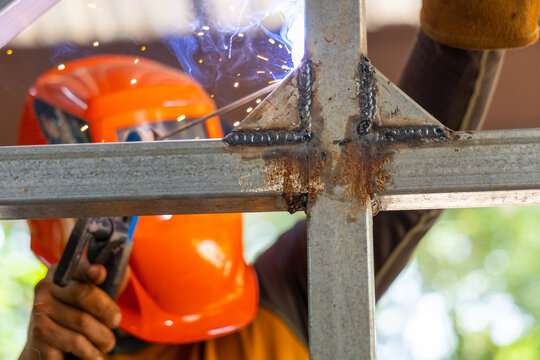 Welder wearing orange helmet welding steel frame joint with sparks and bright arc, closeup showing metal welds, glove and industrial safety gear in outdoor construction setting