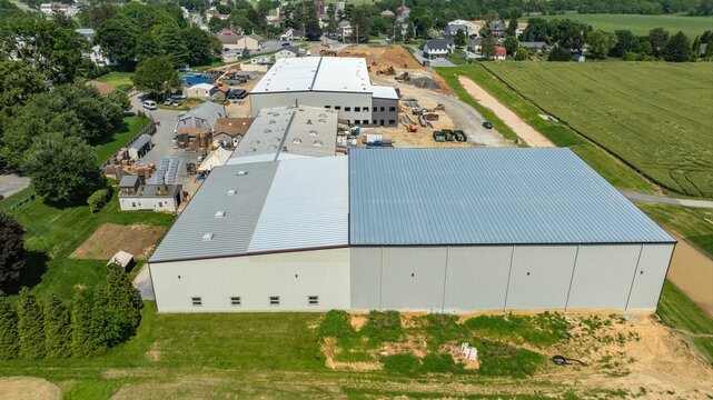 A construction site in a rural area. Large buildings are being built. Nearby, heavy machinery is working on the ground. Trees and fields surround the site.