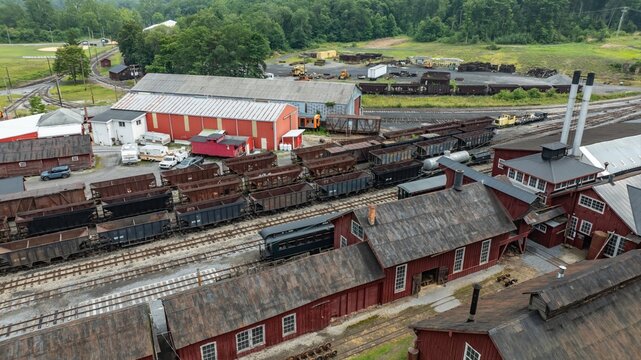 The scene shows a railroad yard with multiple train cars parked on tracks. Old buildings in red and gray surround the yard. Green hills are visible in the background under a clear sky.