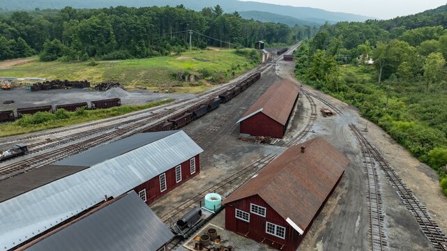 Trains are parked on tracks in an old train yard. Red storage buildings line the area. Green trees cover the hills in the background. The scene shows a quiet, working railway site.