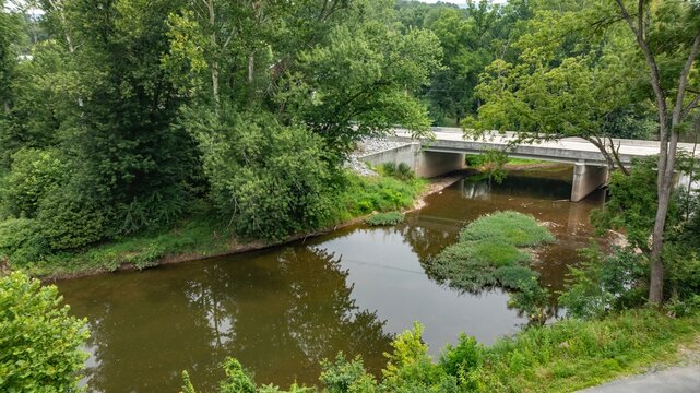 The scene shows a river flowing quietly under a bridge. Green trees line both sides of the river. The water reflects the surroundings. This location is in a natural setting with many plants.