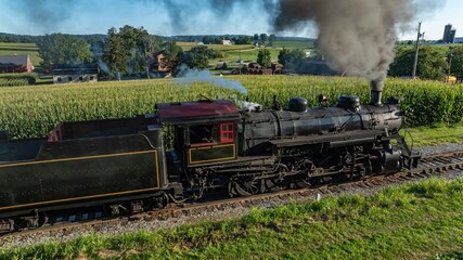 A steam train moves along its tracks, emitting smoke as it passes through fields of tall corn. The train is surrounded by green farms and rural scenery under a clear sky. © Greg Kelton