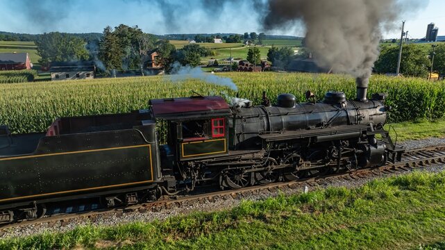 A steam train moves along its tracks, emitting smoke as it passes through fields of tall corn. The train is surrounded by green farms and rural scenery under a clear sky.