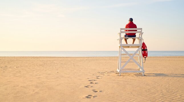 Safety professional sitting on lifeguard tower overlooking calm empty beach and blue ocean waters at morning dawn