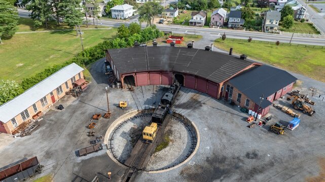 Steam locomotive enters a roundhouse at the train yard. Surrounding buildings show a mix of old and new styles. Grass and road appear nearby in a small town setting under clear skies.