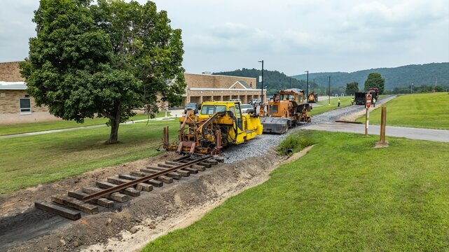 Construction workers are laying down new railroad tracks next to a school. Machines are in operation, and the area is open with grass and trees. The sky is overcast.