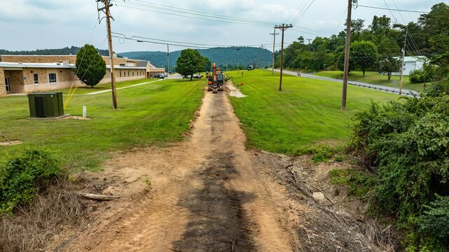 Construction is taking place on a rail road right of way next to a school building. Workers are using machines to clear the area. Power lines run beside the path in a rural setting on a cloudy day.