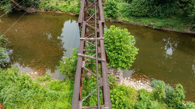 A rusty rail road bridge stands over a river with clear water and visible stones. Green trees frame the area. The scene shows a rural setting with natural vegetation.