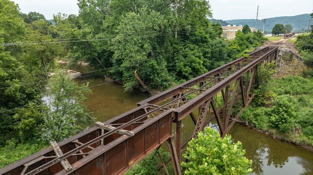 A rusty old abandoned railway bridge spans a river in a rural location. Lush green trees line the banks, and a small path is visible nearby. Power lines run above the bridge during a sunny day.