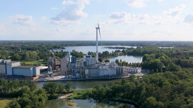 Aerial view of a modern industrial factory plant near a forest and water reservoir with wind turbines. Sustainable manufacturing and green power facility.