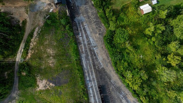 Aerial view of a long set of train tracks next to green trees and small buildings. The scene shows a rural area with some open land and no trains present.
