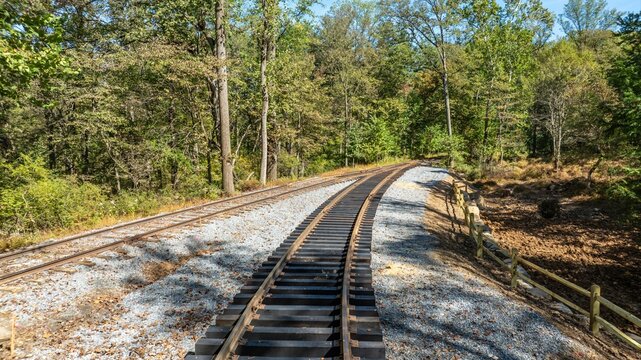 Railroad tracks run along a gravel path in a forested region. Trees surround the area, and sunlight shines on the tracks. The setting shows a rural landscape away from towns.