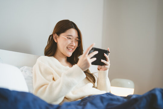 Happy smile young adult asian gamer woman wear eyeglasses play a online game portable handheld in bedroom at night