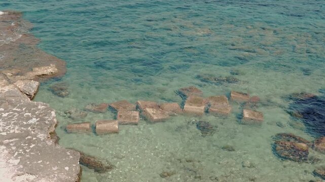 The crystal-clear turquoise waters of Sicily lap gently against the rocks of a cove in Syracuse under a bright sun