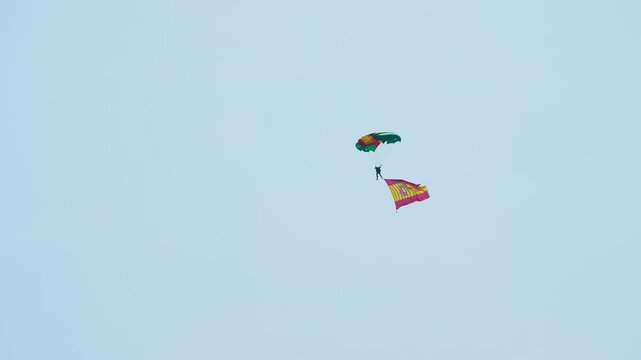 Parachutist descending under a colorful canopy while trailing a large Spanish flag against a pale blue sky, skydiver gliding down during an aerial display demonstration