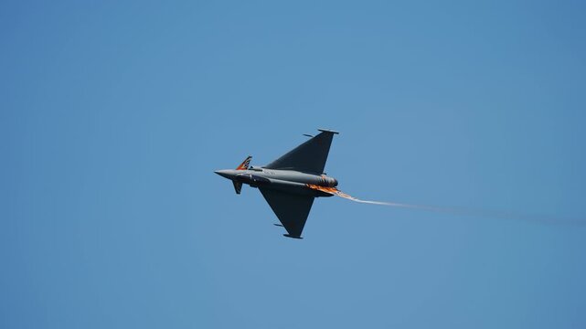 Gray twin engine delta wing canard military fighter jet performs a steep bank with orange afterburner flame visible from engine against cloudless blue sky