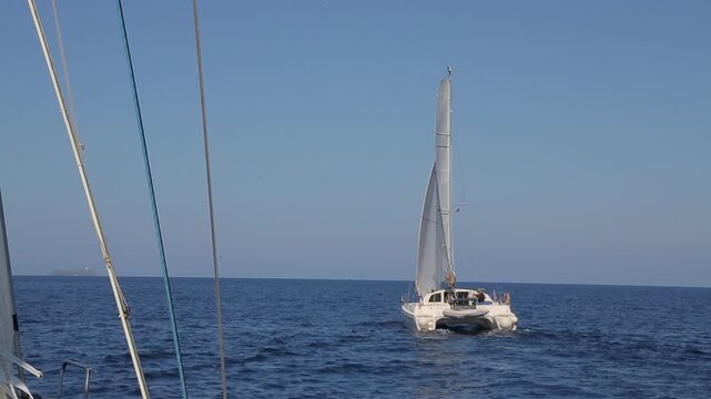 Point of view shot from a sailboat looking at a modern luxury catamaran cruising under full sail on the deep blue calm waters of the Mediterranean Sea. The scene captures the beauty of nautical travel
