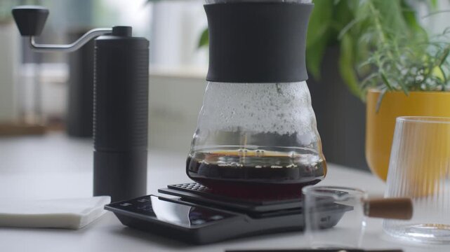 Coffee drips through paper filter into glass server on digital scale beside manual grinder. Concept of pour over brewing and manual coffee preparation