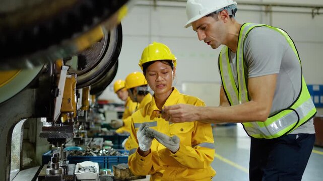 Male factory supervisor in a hard hat inspects a metal part produced by asian female worker operating a heavy industrial stamping machine in a modern manufacturing plant. Teamwork and quality control