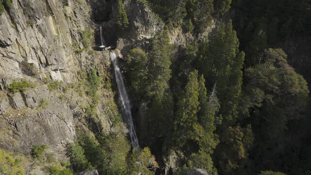 Aerial view of Cascada Los Cesares waterfall cascading down a steep rocky cliff surrounded by lush green pine forest in Nahuel Huapi National Park, Rio Negro, Argentina.