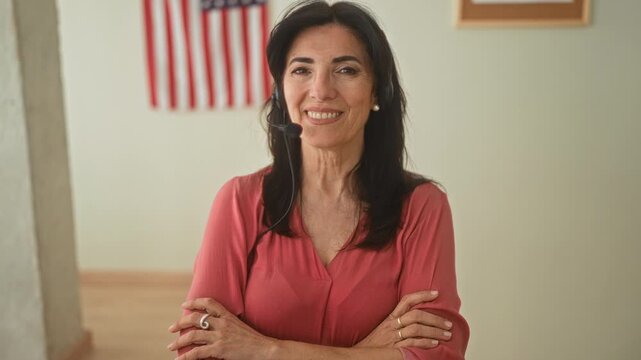 Middle age hispanic woman with headset and arms crossed, hands visible, smiling in a public building with an american flag on the wall; confidence community support.