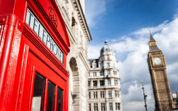 Traditional red telephone box in London. Classic British icon stands near Parliament with Big Ben visible under a bright blue sky.