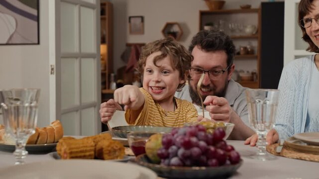 Caucasian kid sitting at dinner table with father and grandmother, pointing at food and smiling. Clip for family lifestyle commercials, healthy eating ads, and parenting promos