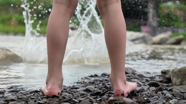 Caucasian child boy stands on stones on the river. Picks up a stone and throws it into the river, splash. Children's legs and feet. Vacation, leisure, rest in a mountain river. Happy childhood