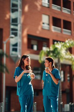 Two female nurses taking a break outdoors, drinking coffee and talking. Colleagues having a conversation about nursing roles