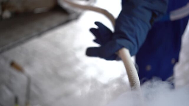 A person wearing protective gloves and clothing handles a hose releasing vaporous liquid nitrogen outdoors.