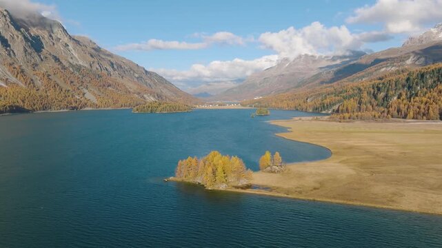 Aerial drone video of mountains and a lake in Engadin valley, Switzerland. Sunny autumn landscape with golden trees and water reflection in motion.