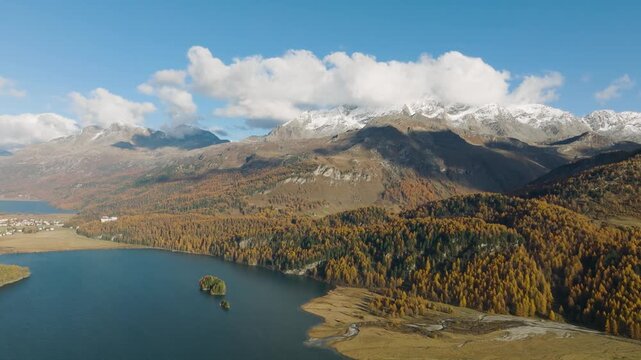 Aerial drone video of mountains and a lake in Engadin valley, Switzerland. Sunny autumn landscape with golden trees and water reflection in motion.