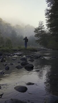 Lone hiker with a backpack stands on the rocky bank of a river in a misty autumn forest, surrounded by tall trees and natural wilderness.